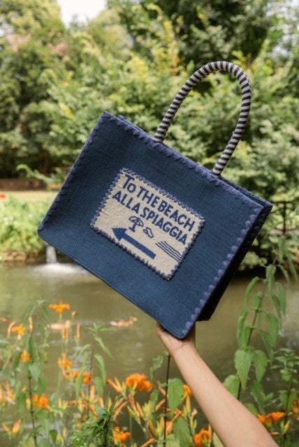 Background beautifully hand crafted designer Navy Blue De Siena jute beach bag, with beaded detail and raffia embroidery along with a cotton lining. Perfect for your next luxury trip to paradise.